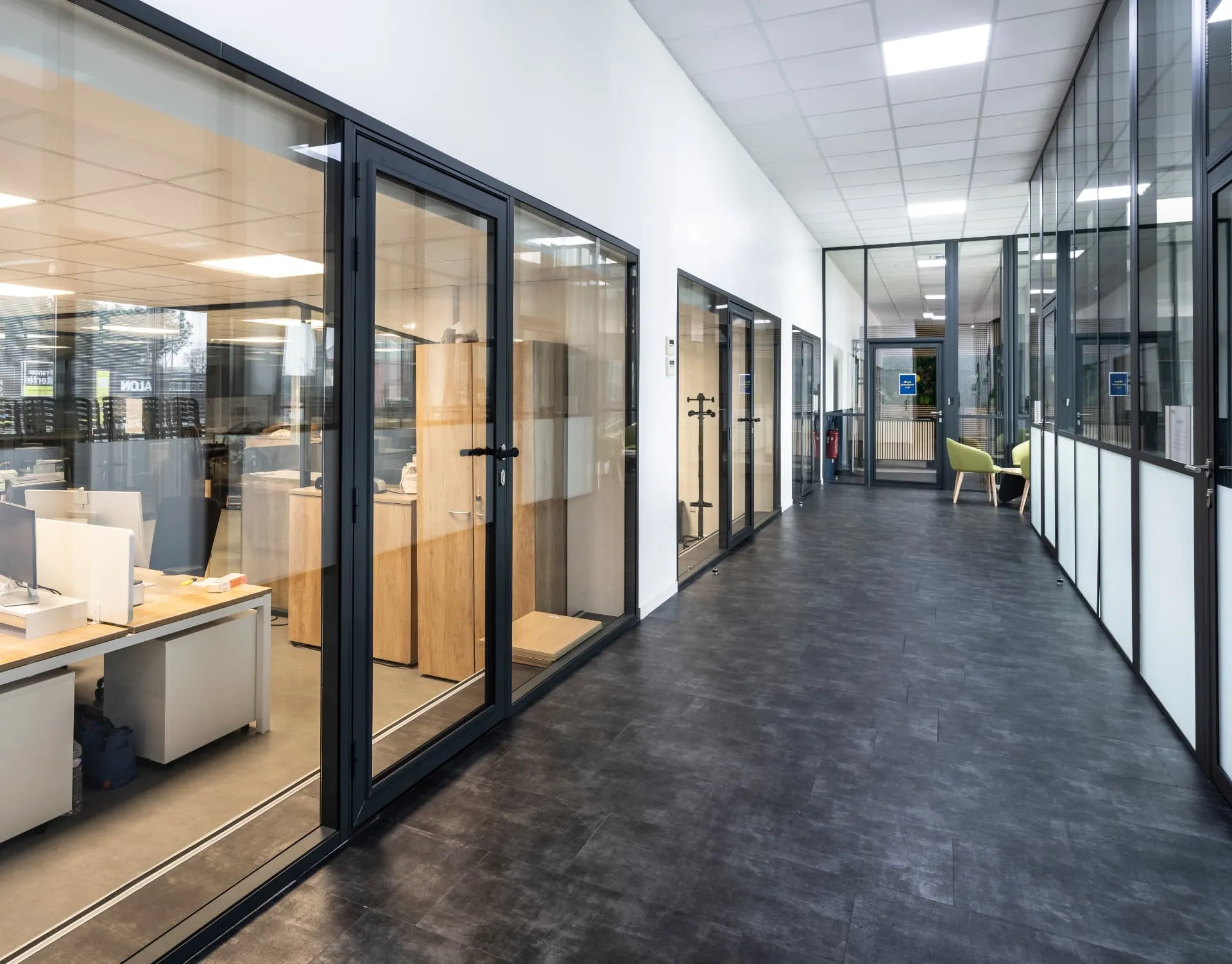 Couloir de bureaux aux cloisons vitrées noires, sol gris foncé et plafond lumineux