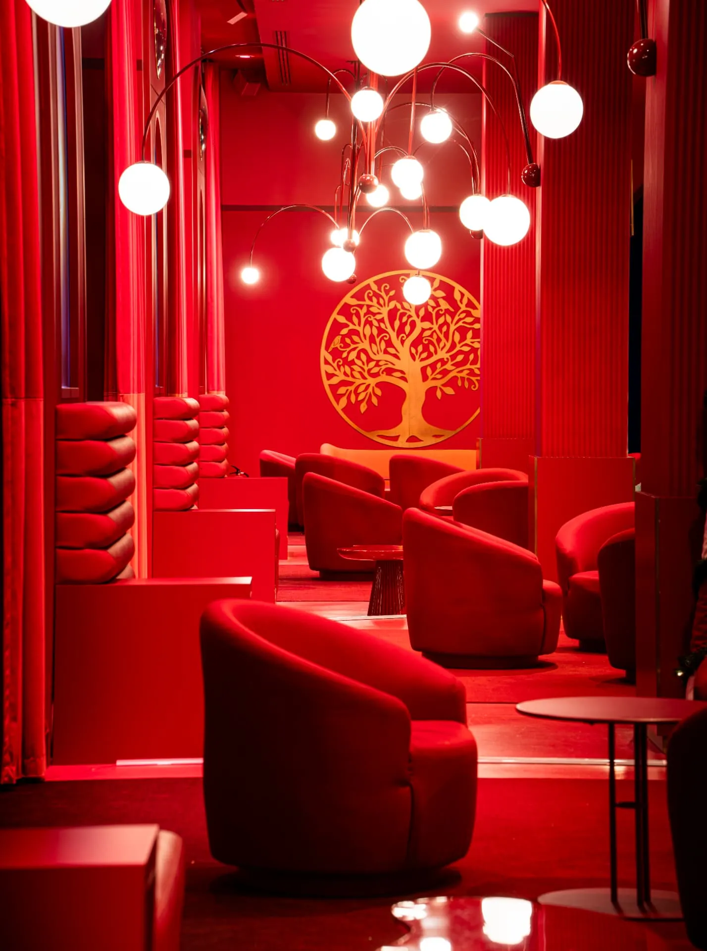 Salon entièrement rouge avec fauteuils en velours, suspensions à globes lumineux et arbre doré au mur