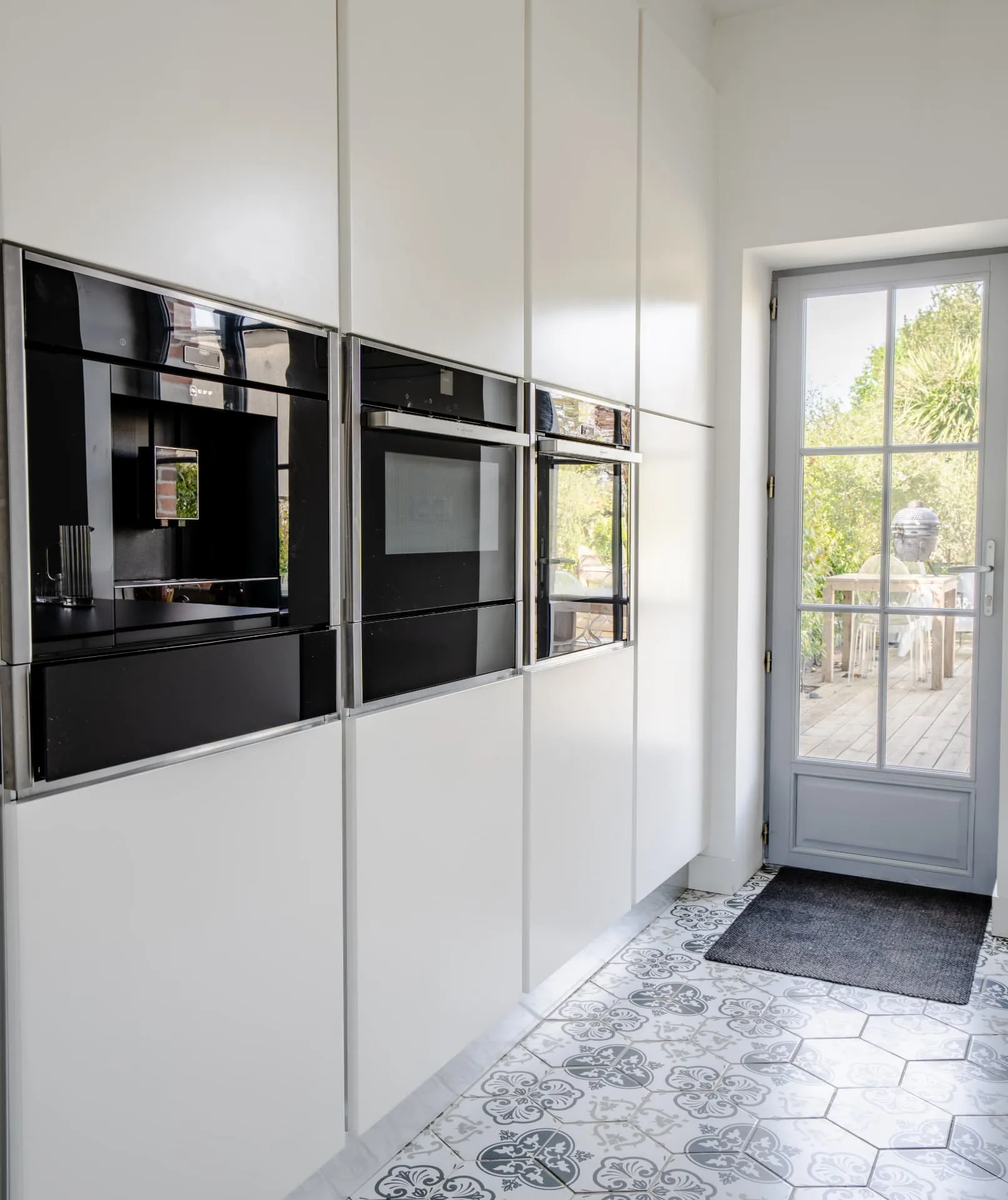 Cuisine blanche aux fours encastrés noirs, carreaux de ciment et porte vitrée ouverte sur une terrasse