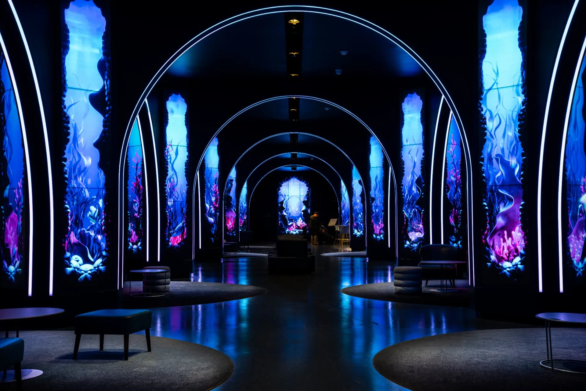 Enfilade d’arches lumineuses encadrant des écrans aux décors sous-marins, sol sombre et reflets bleus