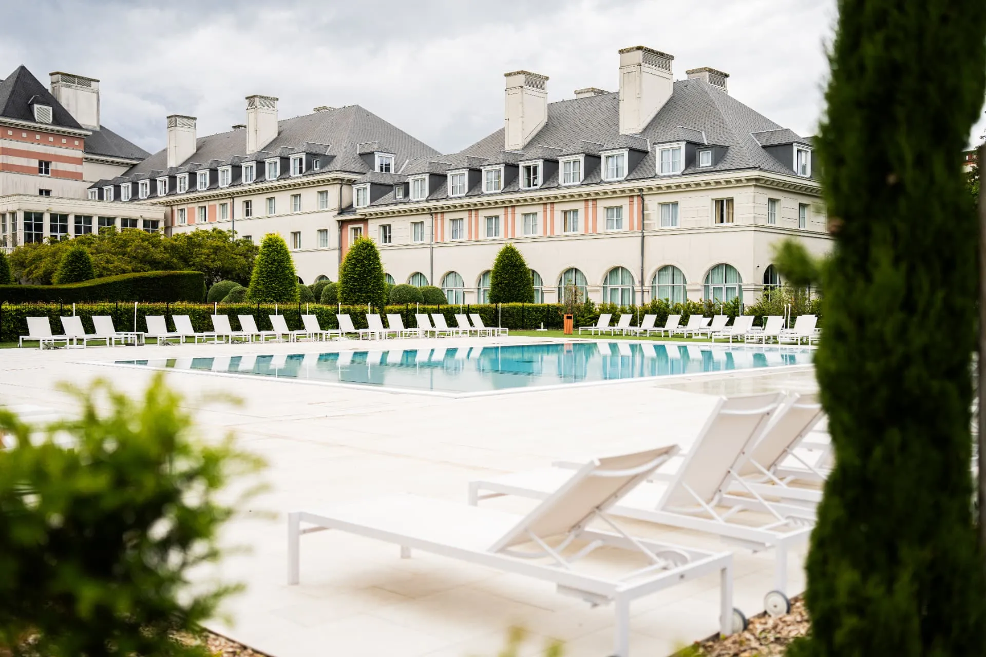 Piscine extérieure bordée de transats blancs devant un grand bâtiment aux toits d’ardoise et lucarnes
